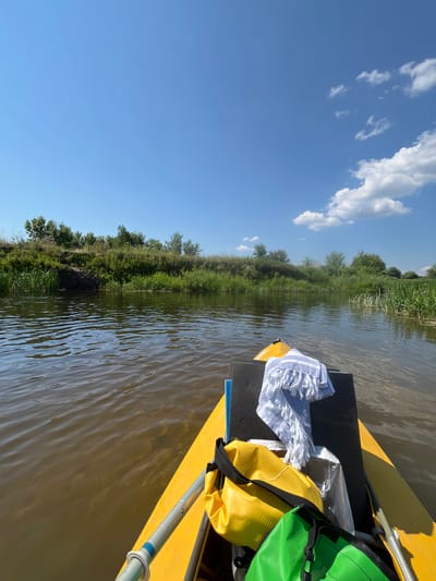 The image shows a first-person perspective from a yellow kayak on a calm, brown river. The riverbanks are lined with lush green vegetation and trees under a clear blue sky with scattered white clouds. In the foreground, on the kayak, are several dry bags: one yellow, one green, and a white and blue striped towel is draped over a black pad. Part of a paddle is visible on the right side. The scene appears to be a serene, natural environment, possibly in Zagorshchino, Russia. There are no people visible in the image.