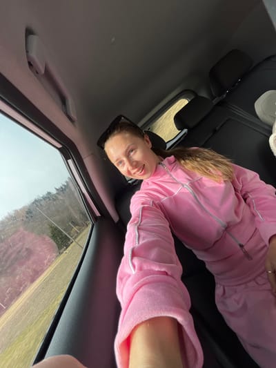 The image displays a female subject seated inside a vehicle, likely in Lugovi, Montenegro. The subject has light brown hair in a ponytail, with black sunglasses resting on top of her head. She is wearing a pink velour tracksuit featuring silver piping details and a full-zip top. Her left arm is extended towards the camera, indicating she is holding the device. A ring is visible on her left hand's ring finger. She is smiling and looking directly at the camera. The vehicle's interior includes dark-colored seats, a light grey headliner, and a white grab handle positioned above a side window. Through the window, an outdoor scene is visible, consisting of a clear sky, a light brown field, and several trees, some bare, suggesting a rural or semi-rural landscape. Vertical utility structures are also present in the background.