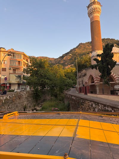 The image shows a view of a street in Bayındırlık Mahallesi, Turkey, with a prominent minaret of a mosque on the right and apartment buildings on the left. A tow truck or flatbed truck is partially visible in the foreground, with its yellow and gray platform taking up the lower portion of the frame. The truck appears to be parked or in the process of loading/unloading.

The street runs alongside a dry creek bed or drainage channel, lined with stone walls. Lush green trees and vegetation grow along the channel and behind the buildings. In the background, a rocky, tree-covered hillside is illuminated by the warm light of what appears to be sunset or sunrise.

On the street, a few individuals are present. A person wearing a headscarf is seated on a scooter on the left side of the road. A silver car is parked further down the street. The apartment building on the left has several balconies and windows. The mosque features distinctive red and white striped arches and a tall, fluted brick minaret. The sky is clear and a light blue.