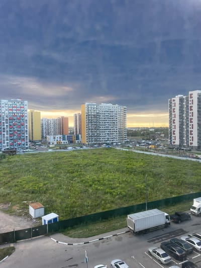 The image depicts a city scene in Domodedovo, Russia, characterized by a mix of residential buildings, green spaces, and infrastructure. In the foreground, a paved area with white and black curbs shows several parked cars, including a silver sedan, a dark sedan, and a white car. A large white truck with a box trailer is also parked on the paved surface. Adjacent to this is a green fence, behind which lies a field of tall green grass, interspersed with some yellow wildflowers. Two small utility structures, one white with blue doors and another with a rusted roof, are visible within the grassy area.

Beyond the grassy field, a row of multi-story residential buildings dominates the middle ground. These buildings vary in color and design, with some featuring colorful balconies or facades. A cluster of smaller, brightly colored buildings that appears to be a school or kindergarten is situated among the residential blocks. Beyond these, more residential buildings and a distant cityscape are visible under a cloudy sky. The sky itself is a dramatic blend of deep blues and grays, with patches of lighter clouds near the horizon, suggesting either sunset or an approaching storm. The overall impression is one of a developing urban area with ample green space, situated in a location identifiable as Domodedovo, Russia.