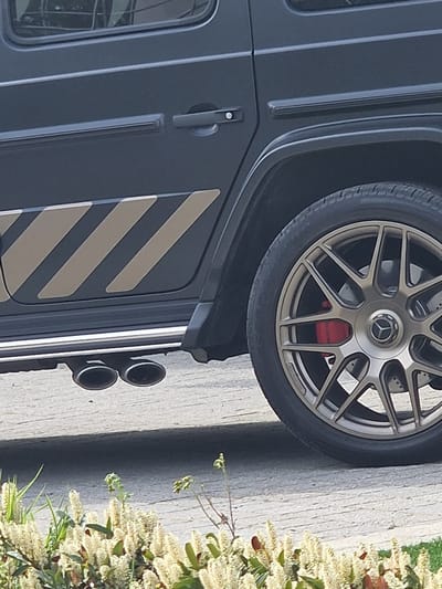 A close-up shot of a dark-colored SUV, presumably a Mercedes-Benz G-Class, is positioned beside a sidewalk in Witton Gilbert, United Kingdom. The SUV features a distinctive wheel with a multi-spoke design and red brake calipers visible behind the wheel. The side of the SUV is adorned with contrasting diagonal stripes and a set of dual exhaust pipes. A shrub with white blossoms is located adjacent to the vehicle. The vehicle and plant are beside a concrete road. The scene is illuminated with soft daylight, indicative of an overcast or diffused lighting condition. No people or other objects are visible. The image is rotated clockwise to orient the landscape vertically.
