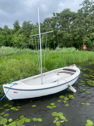 This image depicts a white sailboat moored on a body of water, likely a lake or canal, near a lush green shoreline. The boat, with its mast and rigging, is positioned in the foreground, gently bobbing on the water. Lily pads are scattered across the dark water's surface, with some small yellow flowers visible among them. The background features a dense wall of green trees and reeds, with a cloudy sky overhead. A red and white navigation marker and a wooden structure are visible in the distance on the right side of the frame. The number "481" is affixed to the side of the boat. The scene suggests a serene, natural environment, possibly a park or recreational area. While the prompt mentions Birkerød, Denmark, there are no explicit visual cues in the image to confirm this specific location.