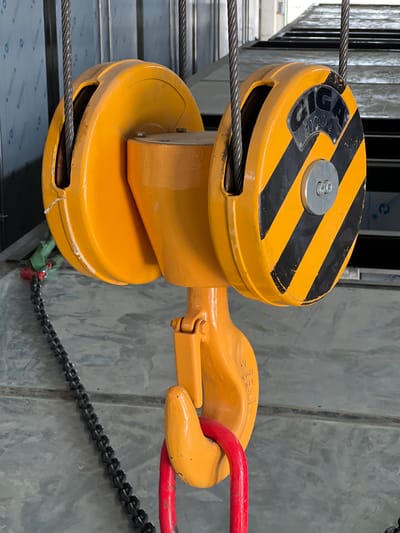 The image shows a close-up, eye-level view of a yellow industrial hoist hook and chain assembly, suspended from steel cables. The hoist hook is large and orange-yellow, with a red circular shackle attached. A black metal chain hangs down from the hook. The hoist mechanism has two large yellow wheels, one with black and yellow hazard stripes and a label reading "GIGA 10,000 kg". The background is a concrete floor and metallic walls or structures, suggesting an industrial or workshop environment. The scene is lit by artificial light. The location is identified as Modřice, Czechia. There are no people or animals visible in the image.