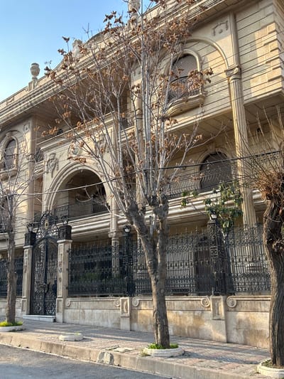 The image captures a grand architectural facade, likely a residential building, in what appears to be Aleppo, Syria. The building is constructed from light-colored stone or stucco, featuring classical elements such as fluted columns, ornate pilasters, detailed cornices, and arched windows. Intricate molding and decorative patterns adorn various parts of the structure. A prominent, dark wrought iron fence with elaborate scrollwork encloses the property along the sidewalk.
Several large, bare-branched deciduous trees line the street in front of the building, suggesting a late autumn or winter season. Some smaller green foliage is visible behind the fence, near the building. A paved sidewalk with a curb runs along the street, separating it from the property. The scene is bathed in bright sunlight under a clear, blue sky, indicating a clear day, likely late morning or early afternoon. No people or visible text are present in the image.