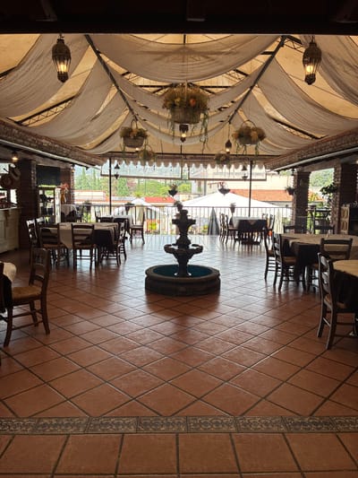 The interior of a restaurant in Tepeji de Ocampo, Mexico, is depicted. A central, tiered fountain sits on a floor tiled in brown squares set diagonally. Tables and chairs, arranged with views of an exterior balcony and beyond, are visible. An arched ceiling with draped white fabric and hanging flower baskets, along with ornate light fixtures, adorns the overhead space. A television is mounted on the wall. The scene appears unoccupied, captured in a well-lit setting.