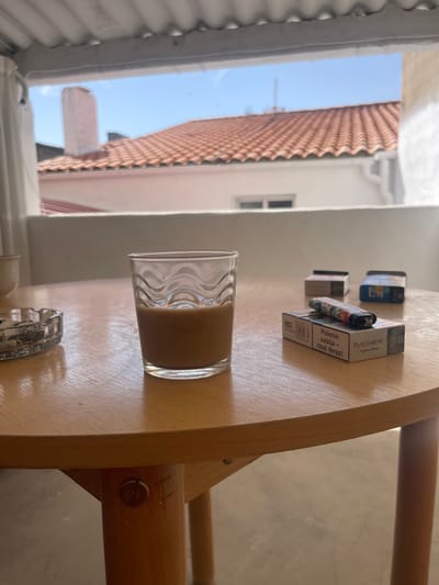 On a wooden table, likely on a balcony in Campillo de Altobuey, Spain, a glass of iced coffee sits next to an ashtray filled with cigarette butts. Two packs of cigarettes and a decorative lighter are arranged to the right of the glass. In the background, terracotta roofs and a chimney are visible under a bright blue sky, suggesting a sunny day. The scene is lit by natural light.