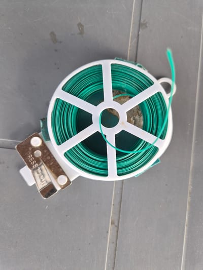 The image shows a close-up, top-down view of a roll of green garden tie wire on a gray, textured surface. The wire is wound tightly around a white plastic spool with a white plastic frame. A small metal lever with the embossed text "PRESS TO CUT" is attached to the bottom of the spool, indicating it's a cutting mechanism for the wire. A single strand of the green wire is visible emerging from the roll and looping towards the top left of the frame. The surface it rests on appears to be outdoors, possibly a deck or patio, given its ribbed texture and a few smudges that could be dirt or water stains. There are no people visible in the image. The lighting suggests it could be daytime, but no specific time or weather conditions can be determined. The context of Lenina, Russia is provided, but there are no visual cues within the image to confirm this specific location. The overall scene is utilitarian, focused on the object and its function.