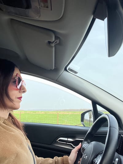 The scene is set inside a vehicle in Olingen, Luxembourg, viewed from the front passenger area towards the driver. An adult individual, identified by dark hair, wearing large pink-framed sunglasses, and red lipstick, occupies the driver's seat. They are dressed in a light brown zippered jacket, with their right hand positioned on the lower rim of a black steering wheel bearing the "KIA" logo. The car's interior features a gray headliner and a raised sun visor. Beyond the driver's side window, an open green field, demarcated by a fence line with wooden posts, extends to the horizon under an overcast sky. A faint line of distant trees is visible beyond the field.