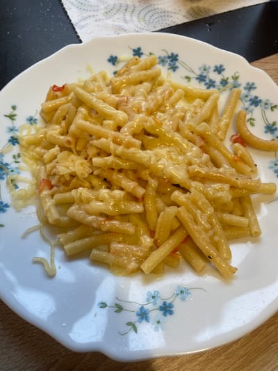 A close-up, overhead shot captures a plate of macaroni and cheese. The pasta, coated in a creamy, melted cheese sauce, is piled high on a white, scalloped-edge plate adorned with a delicate blue floral pattern. Some red pieces, possibly tomato or bell pepper, are interspersed with the pasta. The plate rests on a wooden surface, and to the left, a black surface with a patterned white paper towel is visible. The lighting suggests it's daytime, likely indoors. There is no visible text.
