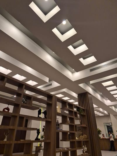 The image shows the interior of a building with a striking geometric ceiling design. The ceiling features angular recessed lighting and square-shaped cutouts. There are also linear vents. In the foreground, a tall wooden shelving unit with various decorative objects is visible. In the background, there is a counter area with people. The location is Boca del Río, Mexico.