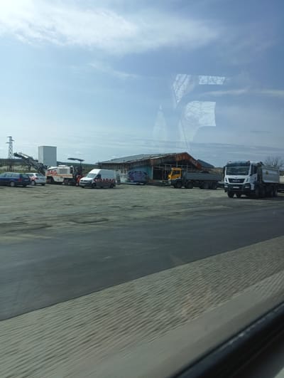 The image shows a view from a moving vehicle, likely a car or bus, towards a construction site in Sokolovo, Bulgaria. The foreground is blurred from the motion. The construction site contains a variety of vehicles and equipment, including an excavator, two MAN trucks, other smaller trucks, vans with "STRABAG" markings, and private cars. Some of the vehicles seem to be parked, while others may be preparing for use. A building with a dilapidated roof is in the background, with faded "Coca-Cola" signage visible. An electrical pylon is on the extreme left. The weather is partly cloudy. The scene suggests ongoing or recently paused construction work in a rural-industrial area.
