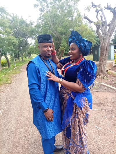 In Tambes Mile Four, Nigeria, a man and a woman are pictured standing on a dirt path. The man, wearing a blue traditional outfit and a black cap, has his right hand on his head. The woman, adorned in a blue velvet dress with elaborate beading and a large blue headwrap, is touching the man's chest. She wears a coral necklace and earrings. The background features lush green trees and foliage, suggesting an outdoor setting with a slightly overcast sky. The overall scene conveys a sense of cultural celebration or a formal occasion.