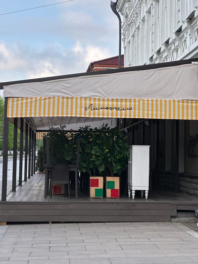 The image shows a cafe awning and outdoor seating area. The awning is white with yellow stripes and has the word "Липовечно" written on it. Below the awning is a plant with lemons. There are chairs and a small table with red and green squares on top. The seating area is on a wooden deck. The background shows a street with a sidewalk. The location is in Akhieli, Georgia.