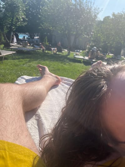 The media file captures a person lying on a towel outdoors, possibly at a resort or park, given the presence of a swimming pool in the background and other people relaxing on lounge chairs. The sun is shining brightly, suggesting a warm, clear day, likely in the late morning or early afternoon. The greenery of a lawn and trees indicate a natural, possibly landscaped, environment. The camera angle is close-up on the person's leg and hair, with the background out of focus. The overall mood seems relaxed and leisurely, characteristic of a sunny day spent outdoors. The specific location is identified as Stăuceni, Moldova.