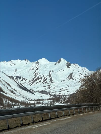 Here is a description of the image:
The image depicts a mountain face covered in snow, seen from a perspective alongside a road. The mountain's snow-covered surface has visible rock features, creating a textured appearance. A clear blue sky fills a large portion of the frame. A section of road with a guardrail is visible on the right, situated between the mountain and the viewer. A line is seen across the sky as well.
Based on the mountainous terrain, snow, and road, the image is likely taken in Le Monêtier-les-Bains, France, given the query context. This area is known for its alpine scenery and proximity to the Écrins National Park.