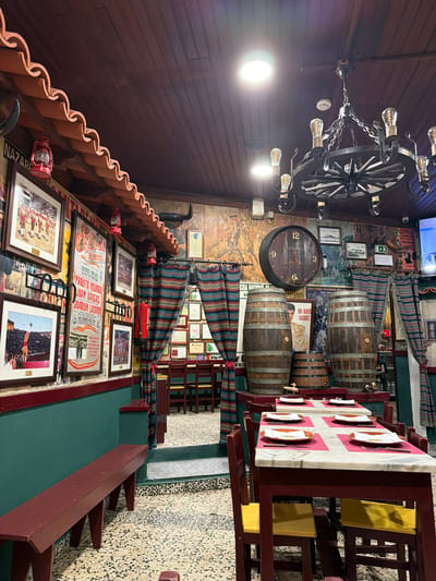 This image depicts the interior of Taberna Quinta Nova, a restaurant located in Santarém, Portugal. The scene presents a traditional, rustic dining environment characterized by bullfighting and equestrian themes.
The ceiling consists of dark brown wooden planks, illuminated by several recessed circular lights and a large, dark metal chandelier designed like a wagon wheel, fitted with multiple exposed filament bulbs. Along the upper left wall, a terracotta-colored, wavy trim resembling roof tiles is visible, beneath which mounted bull horns accent the decor.
The walls are densely decorated. The lower portion of the left wall is painted in a deep teal with a maroon trim. Above this, the wall is covered with framed bullfighting posters (including one for "Praça de Touros Salamanca"), framed photographs depicting bullfighters and arena scenes, and several decorative red lanterns. A red fire extinguisher is mounted on this wall. The background wall features painted murals, additional posters (such as "XXIV Feira Nacional do Cavalo" and one for "Rui Bento Vasquez"), and various framed certificates or documents. A television screen is visible in the upper right.
Two sets of colorful, vertically striped fabric curtains, displaying patterns in blue, red, green, and yellow, frame a stepped entryway leading to a lower dining area at the rear. The floor is composed of light-colored terrazzo with irregular dark speckles.
In the foreground, several dark maroon wooden tables with white marble tops are set. Each table features pink placemats, white rectangular papers, orange plates, and silverware, indicating readiness for patrons. Accompanying the tables are dark maroon wooden chairs, some with yellow seats. A long, maroon wooden bench extends along the lower left wall.
Prominently featured objects include a large, circular wooden wall clock, shaped like a barrel end, displaying "TABERNA QUINTA NOVA" and Roman numerals at 12, 3, 6, and 9. Three wooden barrels of varying sizes, reinforced with metal bands, are positioned on a raised platform behind the foreground tables; one barrel is fitted with a wooden spigot. No people are present in the image.