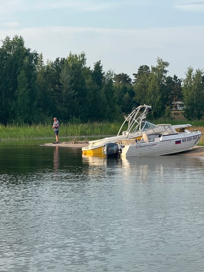 A man stands on a small sandy patch of land in the foreground, fishing. He is wearing a patterned short-sleeved shirt and dark shorts. Two boats are pulled up onto the sand next to him, one is yellow and the other is white with a registration number AA 0261 RUS. The boats are on the shore of a body of water, with a line of green reeds and trees in the background. The sky is a pale blue with some wispy clouds. The location is the city of Borovoye, Russia.