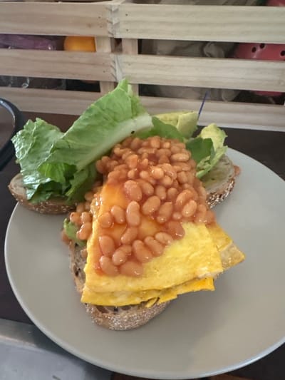 A plate with a sandwich sits on a wooden table. The sandwich is made of two slices of bread, topped with avocado, scrambled eggs, baked beans, and lettuce. In the background, a wooden crate is visible, with various items inside. The scene is indoors, with natural light coming from the left. The plate is light gray, and the sandwich is the main focus of the image. The overall impression is of a casual meal.