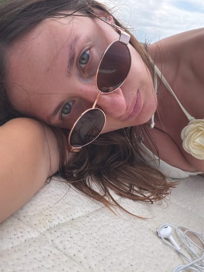 A young woman with brown hair and blue eyes is lying down, resting her head on her arm. She is wearing a white bikini top with a floral embellishment on the strap. Rose gold aviator sunglasses with dark lenses are resting on her face, partially obscuring her eyes. Her lips are slightly parted, and she appears to be looking at the camera. Her hair is long and wavy, and some strands are falling across her face. A pair of white wired earbuds are placed on a textured, light-colored surface next to her. The background is out of focus but suggests an outdoor setting with a cloudy sky. The textured surface she is lying on appears to be a mattress or padded cushion. The specific location of Tsikhisdziri, Georgia is not discernible from the image itself.