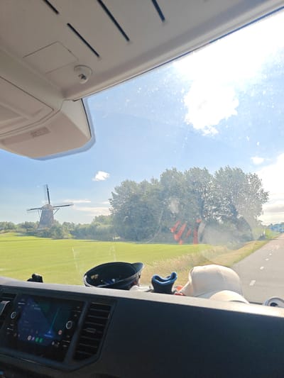 The image captures a view from inside a vehicle, looking out through the windshield on a sunny day. The scene depicts a rural landscape in Leimuiden, Netherlands, with a traditional Dutch windmill visible in the distance. The windmill has dark sails and is situated on a small mound surrounded by lush green fields. A line of trees with full foliage borders the field and extends towards the right side of the image.

In the foreground, the dashboard of the vehicle is visible, with a touchscreen display showing a navigation interface. Various objects are placed on the dashboard, including a black cap with a blue lining, a white baseball cap, and what appears to be a small can. The sky is blue with scattered white clouds, and the sunlight suggests it is daytime, likely mid-morning to late afternoon. The road is visible to the right, with white markings indicating lanes. The overall impression is one of a pleasant, scenic drive through the Dutch countryside. There is no visible text in the image.