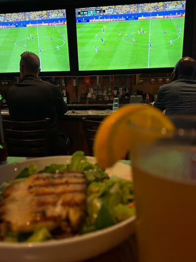 In Riverview, United States of America, a dimly lit bar or restaurant displays a soccer match on two large screens, with the score reading "BRA 0 - 0 HAI" and the time at "22:38". Two men are seated at the bar, facing away from the camera and towards the screens. The man on the left is wearing a black long-sleeved shirt, and the man on the right is wearing a dark patterned shirt. A partially blurred close-up shot of a grilled chicken salad and a glass of orange-colored liquid with an orange slice garnish is in the foreground. The bar area is visible behind the patrons, with bottles of alcohol and various national flags displayed along the back wall.