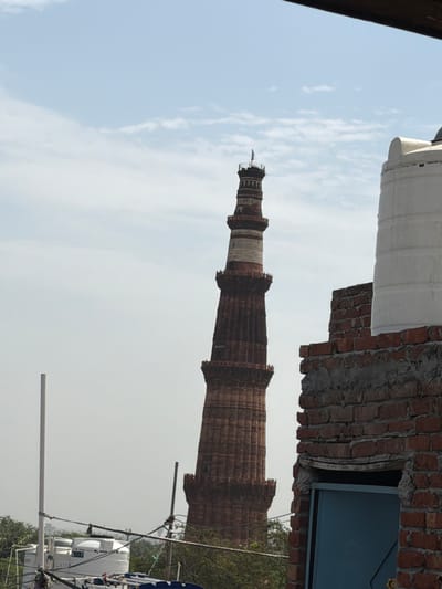 The image shows the Qutub Minar, a UNESCO World Heritage Site, in Delhi, India. The tower, made of red sandstone, is tall and slender with multiple stories. In the foreground, there is a partially visible water tank and a brick structure. The sky is clear with some scattered clouds. The overall scene appears to be a view of the Qutub Minar from a nearby rooftop.