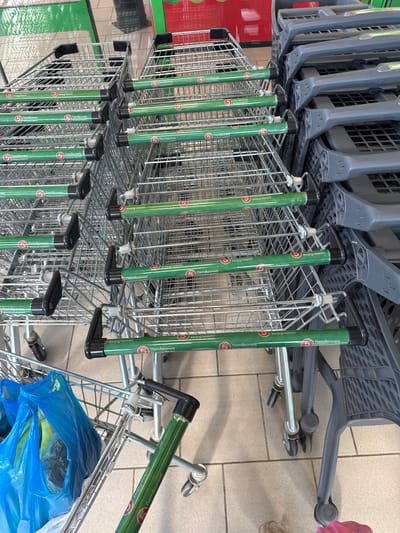 This image shows a row of stacked shopping carts in what appears to be a grocery store or a similar retail environment. The carts are made of metal wire, with green handles that have the "Pyaterochka" logo and text in Russian. Some of the carts are nested inside each other, a common way to store them when not in use. In the foreground, there is a blue plastic bag filled with items, placed inside one of the carts. The floor is tiled. The lighting suggests an indoor setting. The visible text is in Russian and appears to be branding for the "Pyaterochka" supermarket chain. The overall scene depicts a typical retail setting with organized shopping carts. There are no people visible in the image. The image is taken from a slightly elevated angle, looking down at the carts.