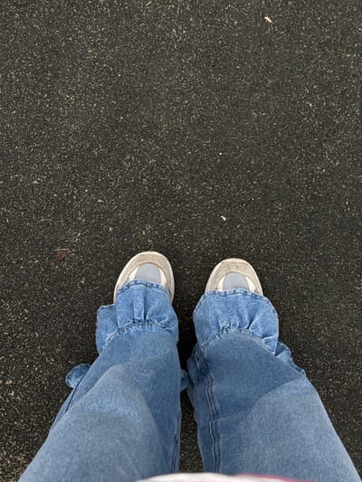 A person is standing on a dark, textured surface. The person is wearing blue denim pants with ruffled cuffs around the ankles and white and grey sneakers. The pants are loose-fitting and gathered at the ankle. The surface appears to be asphalt or a similar material. The image is taken from a high angle, looking down at the person's feet. There are no other people or objects visible in the image. There is no information in the image to suggest it is located in Bylovo, Russia.
