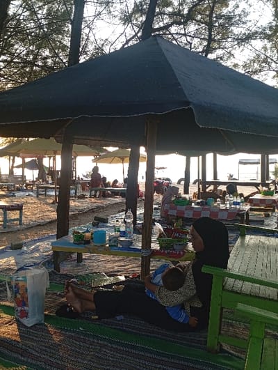 The image depicts a beach scene in Tritunggal, Indonesia, during late afternoon or early evening. The low sun creates backlighting and casts long shadows across the sandy ground. A canopy of slender tree trunks and branches, consistent with pine trees, extends across the upper portion of the frame.
In the immediate foreground, a woman, wearing a black hijab and dark clothing, sits barefoot on a striped mat (green, brown, black). She holds an infant, dressed in a light-colored patterned top and blue garment, who appears to be resting in her lap. To her right, a partial green wooden bench structure is visible.
Beneath a large, dark-colored, hexagonal tarpaulin shelter, supported by wooden poles, a low, light-colored table with blue and yellow accents is positioned. This table holds multiple plastic bottles, including a large clear mineral water bottle, various bowls, cups, and a green plastic basket containing items. Further into the background, under additional lighter-colored tarpaulin shelters, numerous individuals are seated at similar low tables, one of which is draped with a red and white checkered cloth. The sandy beach extends towards a body of water in the distance, where indistinct structures are partially visible. The scene collectively represents a casual outdoor dining or leisure area established on a beach.