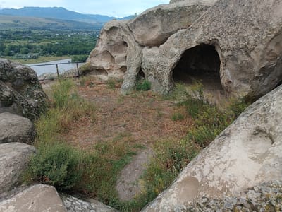 This is a wide shot of Uplistsikhe, Georgia, a cave city. The image shows a cluster of natural rock formations with several cave entrances, some appearing to be carved out. The foreground is dominated by the rough, textured surfaces of the rocks, with sparse, dry grasses and green vegetation growing in patches. A narrow, dirt path is visible winding through the vegetation. In the mid-ground, there is a metal railing that overlooks a valley. Beyond the valley, a river flows, and on the far side, there are green trees and a small town with buildings. The background consists of rolling hills and mountains under a cloudy sky. The overall scene depicts an ancient historical site with a natural landscape.