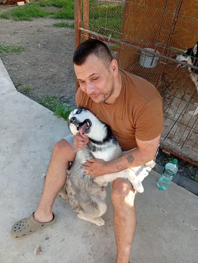 A man sits on concrete, holding a Siberian husky in his lap. The man is wearing a brown t-shirt and grey shorts, and a pair of tan croc-style shoes. The husky is black and white, with blue eyes, and appears to be panting with its tongue out. The man is looking down at the dog with a slight smile. Behind them is a wire-mesh dog kennel. In the background, there is green grass and dirt. A plastic bottle of water rests on the concrete to the right of the man. The location is Podvirne, Ukraine.