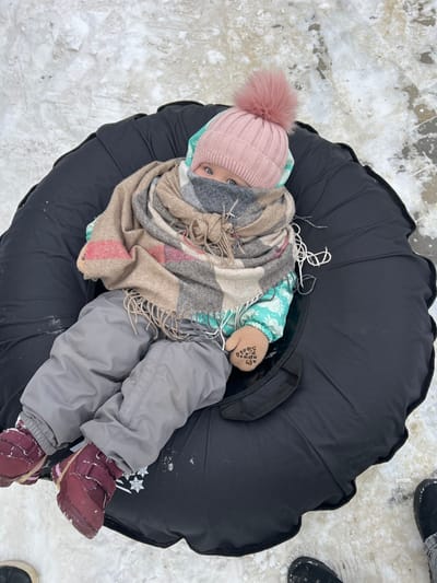 An infant or toddler, heavily bundled in winter attire, is observed sitting upright within a black inflatable snow tube on snow-covered ground in Primorka, Russia. The child wears a light pink ribbed knit beanie with a large pink pom-pom, a multi-colored plaid scarf (tan, brown, grey, red) wrapped around the neck and lower face, obscuring the mouth and chin, and a light blue patterned winter jacket. Grey snow pants and dark red boots with Velcro fasteners cover the lower extremities. Tan mittens with dark grip dots are visible on the hands. The child's blue eyes are visible above the scarf. The immediate environment consists of patchy snow and ice. The toes of dark footwear, presumably belonging to an adult, are visible in the lower left and right corners of the frame, suggesting proximity to another individual.