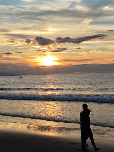 This is a full shot of a beach at sunset in Uleekareung, Indonesia. The sky is filled with soft, wispy clouds painted in shades of orange, yellow, and blue. The sun is a bright orb near the horizon, casting a warm glow over the scene. The ocean is calm, with gentle waves lapping at the shore. Several small boats are visible in the distance. In the foreground, two silhouetted figures walk along the wet sand, their reflections visible in the water. The overall mood is peaceful and serene.