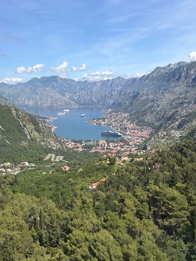 This media file presents a wide, high-angle panoramic view of a scenic bay or fjord surrounded by towering mountains under a bright daytime sky. The scene is dominated by a large body of water, likely a bay, nestled between steep, rocky mountains on the left and densely forested slopes on the right.
In the center of the image, a vibrant town or city with numerous buildings, characterized by their reddish-orange tiled roofs, stretches along the water's edge and extends up the lower mountain slopes. A large white cruise ship is prominently visible, either docked or anchored in the bay near the town, alongside several other smaller boats.
The mountains on the left side appear rugged and craggy, with patches of sparse vegetation, receding into the hazy distance. On the right, the mountainsides are covered in lush, dense green forests. A distinct feature on the forested slope is a winding road with multiple hairpin turns, indicating a challenging mountain pass. Scattered houses with red roofs can be seen nestled within these forested areas.
The sky is a clear, bright blue with scattered white, puffy cumulus clouds. The lighting suggests it is mid-day, with good visibility. No specific human activities are discernable due to the vast scale, but the presence of the town and ships indicates a lively area. There is no visible text in the image. The overall impression is one of majestic natural beauty combined with historical urban development, strongly indicative of the Bay of Kotor region in Montenegro, consistent with the Čavori viewpoint.