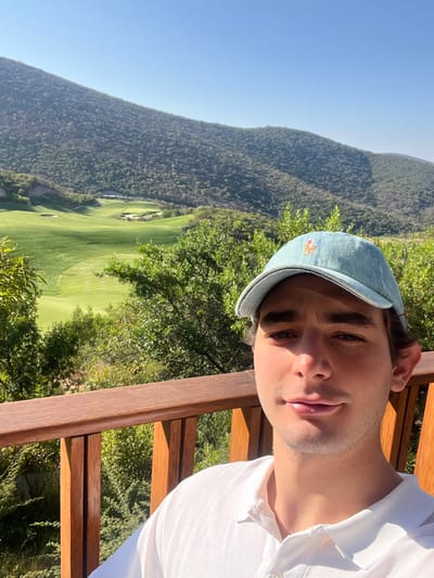 A young man takes a selfie with a scenic mountainous landscape and a golf course in the background. The image is oriented vertically (portrait), with the man's head and upper torso filling the right side of the frame. He is looking directly at the camera with a calm expression, his lips slightly parted. He has dark hair, dark eyes, and a light five o'clock shadow.
The man wears a light blue or denim-colored baseball cap with a red and blue embroidered logo (possibly a polo player on a horse) on the front. He is also wearing a white polo shirt with a collar and buttons. His left shoulder and arm are visible. He is positioned next to a wooden railing, which runs horizontally across the mid-right of the image, suggesting he is on a balcony, deck, or viewing platform.
Behind him, a vast, lush green mountain covered in dense trees dominates the left and center background. Below the mountain, a sprawling green golf course with several fairways and sand traps is visible. The sky above is clear and bright blue, indicating sunny weather. Strong sunlight casts distinct shadows, illuminating one side of the man's face and highlighting the greens of the landscape. The scene evokes a tranquil, outdoor setting during daytime. Given the location context, this is San Isidro Huilotepec, Mexico.