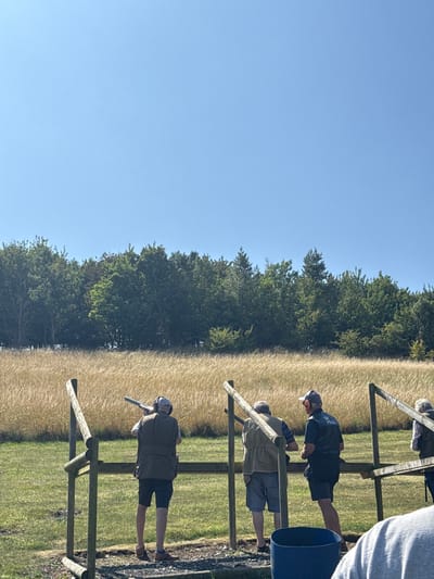 The image displays an outdoor clay pigeon shooting range located in Wold Newton, United Kingdom, under a clear, cloudless blue sky. Four adult males are visible. In the left foreground, an individual wearing a light-colored cap, ear protection, and a dark vest stands within a wooden shooting stand, shouldering a shotgun and aiming it towards the field. To his right, a second male in a light vest stands observing. Further right, a third male, wearing a dark cap and orange ear protection, stands with his back partially turned, observing. A fourth male is partially visible on the far right. The immediate ground consists of green grass and a dark, possibly gravel, surface around the shooting positions. Beyond the foreground, a large field of tall, golden-brown grasses extends towards a dense treeline that borders the horizon.