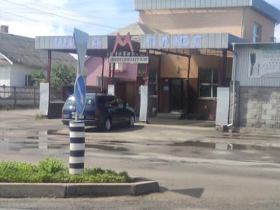 A street-level, eye-level outdoor shot captures a business with a sign overhead that reads "ШИНА ПЛЮС" which translates to "TIRE PLUS." Below this, another sign says "ШИНОМОНТАЖ," meaning "TIRE FITTING." This establishment is located in Kostopil, Ukraine. The building has an orange-brown facade with a white trim, and a light beige colored roof. A dark blue car is parked in front of the business. The ground is wet, with puddles reflecting the sky and the surrounding environment. A striped bollard, black and white, stands in the foreground to the right of the blue car, and a blue directional sign is mounted on top of it. Green grass and foliage are visible along the curb in the foreground. The sky is partly cloudy.