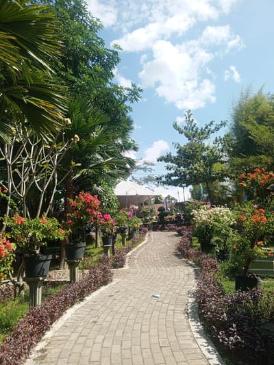 The image depicts a paved pathway winding through a lush garden. Palm trees and other foliage frame the pathway, with potted flowering plants lining its edges. In the background, a white tent structure is visible, suggesting an outdoor event or venue. The sky above is a clear blue with scattered white clouds. The scene appears to be a well-maintained garden, possibly part of a public space or a private residence. There are no people or animals visible in the image. The location is identified as Darsono, Indonesia.