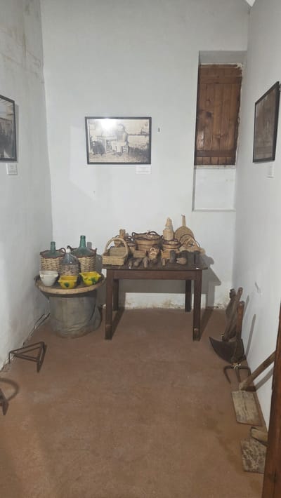The image captures a scene in Altura, Spain. The interior space appears to be a historical exhibit or a museum display, with whitewashed walls and a reddish-brown earthen floor. A wooden-framed picture, possibly a black-and-white photograph, hangs on the wall.

A variety of objects are arranged in the room: woven baskets and glass bottles, some encased in woven material, sit on the floor and a dark wooden table. Several other wooden objects with metal implements, possibly farming or crafting tools, lean against the wall in the background. The overall impression is that of a curated display of traditional rural artifacts.