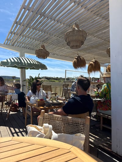 The image depicts an outdoor dining scene at what appears to be a restaurant or bar in Ramatuelle, France, based on the coastal vegetation in the background and the architectural style. Several people are seated at wooden tables, with some individuals in the foreground and others further back. The diners are engaged in conversation or enjoying their meals. A pergola structure with wooden slats covers the area, providing shade. Decorative straw light fixtures hang from the pergola. The flooring is wooden decking. Green and white striped umbrellas provide additional shade. The scene suggests a relaxed, sunlit, and upscale dining experience.