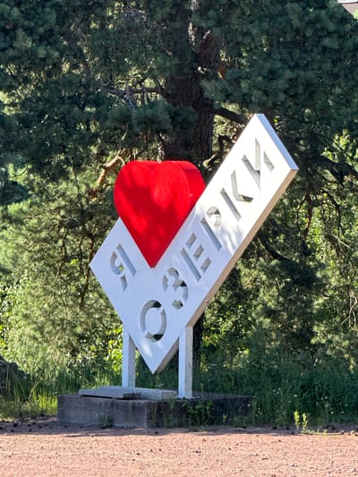 In St. Ozerki, Russia, a white sign with cut-out letters and a red heart is situated outdoors. The sign is mounted on a concrete base and appears to be a landmark. The text on the sign is in Cyrillic and reads "Я ОЗЕРКИ," which translates to "I [heart] Ozerki." The background consists of lush green trees and foliage, suggesting a natural or park-like setting. The ground in the foreground is covered with red gravel. The lighting indicates it is a sunny day. There are no people visible in the image.