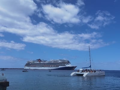 The scene is a waterfront in Cozumel, Mexico, located at 20.512 latitude, -86.95 longitude. A large cruise ship is docked at the pier, its hull painted in white and blue with a large red logo. A white catamaran is sailing past the cruise ship, its white sails are fully extended, and it is marked "Jolly Roger" on the stern. The water is calm and blue, and there are a few small boats in the distance. The sky is blue and clear with a few white clouds. Several people are visible on the catamaran, and they appear to be enjoying the sunshine and the view. The scene is peaceful and serene.