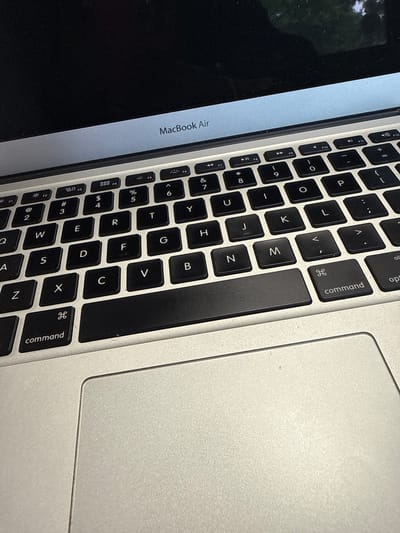 The image shows a partial view of a MacBook Air, likely taken indoors given the even lighting. The focus is on the keyboard and trackpad area of the laptop. The keys are black with white lettering, and the trackpad is a silver-grey color. The screen is visible to the left, showing a dark reflection of what appears to be greenery. The brand "MacBook Air" is printed on the casing near the screen.
Since the image contains a laptop, with no evidence to the contrary, it is impossible to determine any information regarding the location of the laptop. There are no details to infer the presence of Loganville, United States of America.
