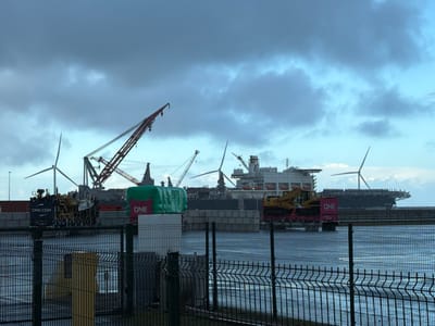 An outdoor wide shot captures an industrial port scene at Maasvlakte, Rotterdam, Netherlands, under a predominantly overcast sky with patches of lighter blue. In the immediate foreground, a dark gray metal security fence runs horizontally across the frame, partially obscuring a calm body of water beyond it.

The mid-ground features a concrete or paved dock area along the water's edge. Multiple shipping containers are visible, including a green, tarp-covered container marked "ONE" and several black containers marked "CMA CGM." A large, multi-wheeled piece of heavy machinery, possibly for container handling, is positioned near the "CMA CGM" containers. Further to the right, a yellow excavator is loaded onto a pink flatbed trailer, also marked "ONE."

In the background, several large vessels and structures dominate the horizon. A massive heavy-lift vessel, identified as the *Pioneering Spirit* by its distinctive design and numerous lifeboats along its broad hull, is docked. Above and around the vessels, multiple industrial cranes with red and dark gray gantries extend into the sky. Additionally, three prominent wind turbines are visible, their blades either stationary or slowly rotating. No individuals or explicit actions are discernible within the frame.