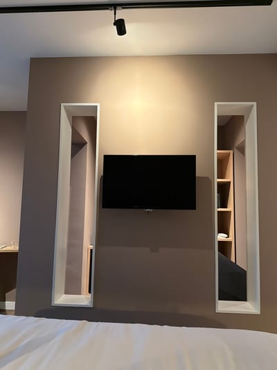 This is a photograph of a wall in a room, likely a hotel room, in Otterlo, Netherlands. A black television is mounted on the wall. To the left and right of the television are tall, narrow recessed shelves, each with a white frame. The wall is painted a light brown color. Above the television, a track light fixture is visible on the ceiling. To the far left, a small table with a glass on it is partially visible. On the right, within the recessed shelving, is a wooden shelf unit with several shelves. The foreground of the image shows a white duvet on a bed. The overall impression is one of modern, minimalist decor.