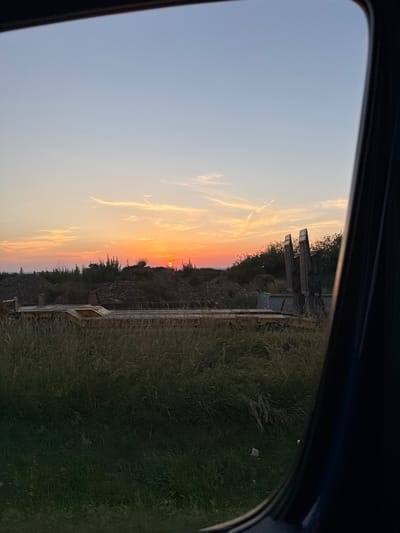 The image depicts a sunset viewed through a car window. The sun, a small orange orb, is low on the horizon, casting a warm glow across the sky. Wispy clouds streak across the sky, illuminated by the setting sun in shades of orange, pink, and yellow. The foreground is dominated by a patch of dry grass, suggesting a natural, possibly rural setting. In the mid-ground, there are some man-made structures, including what appears to be a small, weathered building or shelter with angled roofs. There is also a long, narrow structure, possibly a bridge or walkway, running horizontally through the scene. The overall impression is one of tranquility and the end of a day. The time of day is clearly sunset. There is no visible text in the image. The location context is Oberwetz, Germany, which aligns with the appearance of the landscape.