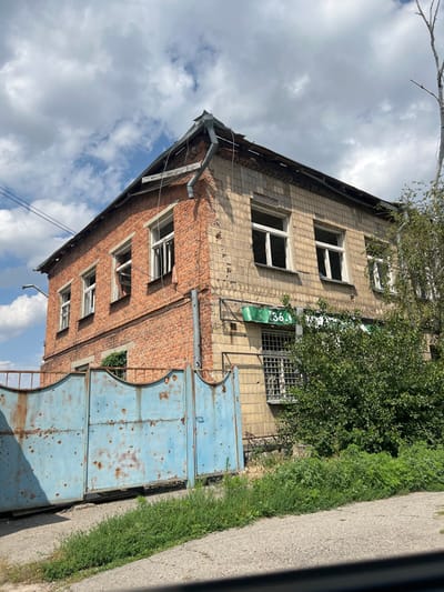 The image depicts a two-story building exhibiting significant damage, located in Staryi Saltiv, Ukraine, under a partly cloudy sky. The building's left facade is constructed of red brick, while its right facade is faced with light-colored, square tiles. The roof structure is visibly compromised, with missing sections of roofing material and a bent metal downspout at the corner. All discernible windows are broken, featuring missing glass panes and damaged frames. The ground-level windows on the tiled facade are fitted with metal security bars. A green sign displaying the number "36-1" is affixed to the tiled facade. In the foreground, a large, light-blue, two-panel metal gate with substantial rust and numerous circular indentations, consistent with projectile impacts, is positioned on an asphalt surface. Green vegetation, including a large bush, is present around the building's base and in the foreground, with a bare tree branch visible against the sky on the right side. No individuals or specific actions are depicted.