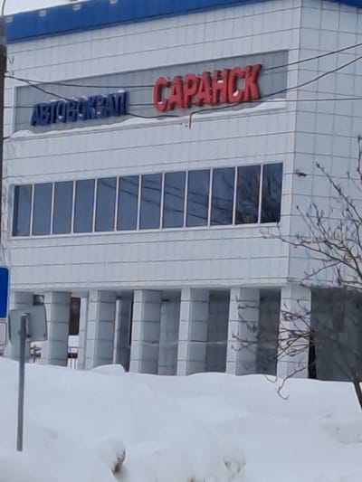 A tilted eye-level shot captures a multi-story building with "CAPAHCK" vertically displayed in red lettering. Beneath it, "АВТОКРАЙ" appears in blue. The building features a line of narrow, tall windows. Adjacent to the building, a gray structure of horizontal levels is visible, partially obscured by a large snowdrift. Bare tree branches are seen above the building.