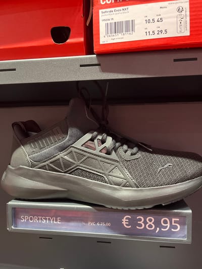 A dark grey Puma Softride Enzo NXT sneaker is displayed on a grey shelf in a retail environment. The sneaker is positioned diagonally, with its toe pointing to the right. A price tag in front of the shoe displays "SPORTSTYLE" and the price "€38,95". Behind the shoe, a red Puma shoebox with a label indicating size information is visible. The label shows "Softride Enzo NXT puma black-pumapuma black" and various size conversions, including UK 10.5, EUR 45, US 11.5, and CM 29.5. The background consists of a dark, blurred surface, likely shelves or walls of a store. The lighting is even, suggesting indoor retail illumination. The scene is static and does not contain any people. This retail display is located in Rodengo, Italy.