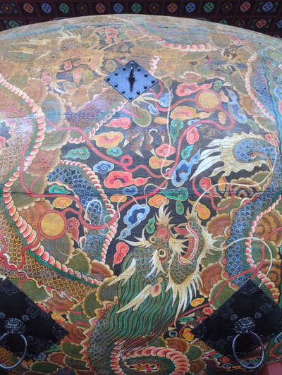 This image presents a close-up, upward-angled view of a large, curved, wooden surface, identified as a traditional Korean drum (beopgo), located in Godang-ri, South Korea. The drum is extensively decorated with vibrant, traditional polychrome artwork.

The primary subjects of the artwork are multiple stylized dragons. Their bodies are rendered with intricate scales in shades of blue, green, and gold, accented with red. The dragons coil and intertwine across a deep black background. Their heads feature prominent eyes, sharp facial features, and flowing manes. One dragon's head is clearly visible in the lower right, another partially visible in the lower left, and a faded representation in the upper left. Interspersed among the dragon forms are numerous stylized cloud motifs in various colors, including red, blue, green, orange, and yellow. Several golden orbs, or "cintamani" pearls, are strategically placed within the composition, notably one in the lower-middle left and a faded one in the upper left.

Affixed to the drum's surface are three distinct pieces of black metal hardware. The uppermost piece is a diamond-shaped plate secured by four rivets, featuring a single, upward-curving metal loop or handle. The two lower pieces are also diamond-shaped plates, each adorned with a sculpted lion's head or beast's head from which a circular metal ring handle is suspended. These plates are also secured with decorative studs.

The paintwork, while still vibrant, shows signs of age and weathering, particularly in the upper sections where colors appear somewhat faded and the underlying wood texture is more visible.

In the uppermost background, a section of a dark red-brown architectural element is visible, likely an eave or ceiling beam of a traditional Korean building. This element features a repeating pattern of square panels, each containing a distinct, stylized floral motif rendered in multiple colors, including red, blue, green, and yellow. No people are present in the scene, and no actions are depicted.